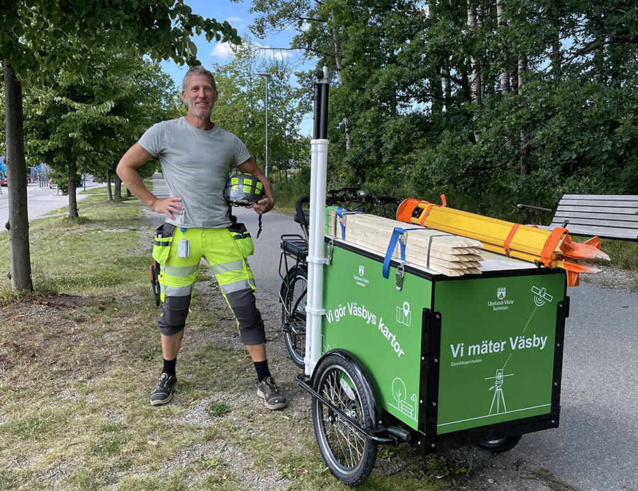 Lådcykel med texten "Vi mäter Väsby" och "Vi gör Väsbys kartor". Bredvid cykeln står en man i arbetskläder.