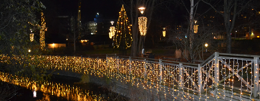Ljusslingor som sitter på räcket längs en å och runt en julgran lyser i mörkret. 