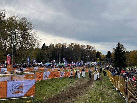 Publik och funktionärer samlade vid målgången under orienteringstävlingen 25-manna i Runbyskogen, omgivna av träd och tävlingsflaggor.